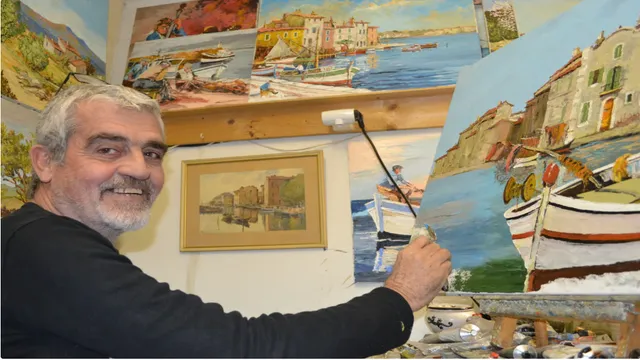 Jacky Buravant souriant, pinceau en main, devant une toile de barques et de maisons au bord de l'eau ; les murs de l'atelier sont couverts de tableaux.